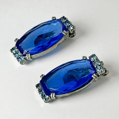 Vintage clip-on earrings featuring large cobalt blue oval rhinestones with light blue chaton accents in silver-tone Art Deco-style settings, mid-century design.
