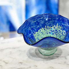 Signed BAGI art glass bowl with a footed base and wavy rim, featuring mottled blue and green swirls. The glass has a glossy finish and sculptural form, with vibrant oceanic and forest tones. Dimensions approximately 5–5½" tall and 7–8" wide.