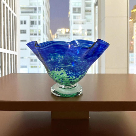 Signed BAGI art glass bowl with a footed base and wavy rim, featuring mottled blue and green swirls. The glass has a glossy finish and sculptural form, with vibrant oceanic and forest tones. Dimensions approximately 5–5½" tall and 7–8" wide.
