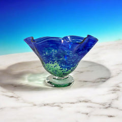 Signed BAGI art glass bowl with a footed base and wavy rim, featuring mottled blue and green swirls. The glass has a glossy finish and sculptural form, with vibrant oceanic and forest tones. Dimensions approximately 5–5½" tall and 7–8" wide.