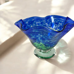 Signed BAGI art glass bowl with a footed base and wavy rim, featuring mottled blue and green swirls. The glass has a glossy finish and sculptural form, with vibrant oceanic and forest tones. Dimensions approximately 5–5½" tall and 7–8" wide.