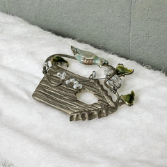 Vintage 1990s silver tone brooch featuring a light blue enamel bird perched on a wire branch with green enamel leaves and white seed bead flowers, set in front of a textured birdhouse; whimsical 3D design in very good vintage condition.