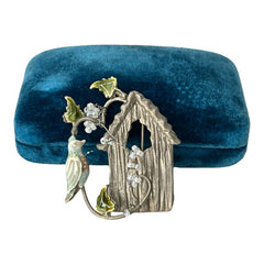 Vintage 1990s silver tone brooch featuring a light blue enamel bird perched on a wire branch with green enamel leaves and white seed bead flowers, set in front of a textured birdhouse; whimsical 3D design in very good vintage condition.