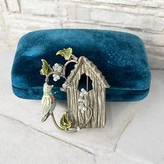 Vintage 1990s silver tone brooch featuring a light blue enamel bird perched on a wire branch with green enamel leaves and white seed bead flowers, set in front of a textured birdhouse; whimsical 3D design in very good vintage condition.