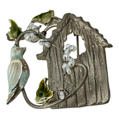 Vintage 1990s silver tone brooch featuring a light blue enamel bird perched on a wire branch with green enamel leaves and white seed bead flowers, set in front of a textured birdhouse; whimsical 3D design in very good vintage condition.