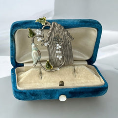 Vintage 1990s silver tone brooch featuring a light blue enamel bird perched on a wire branch with green enamel leaves and white seed bead flowers, set in front of a textured birdhouse; whimsical 3D design in very good vintage condition.