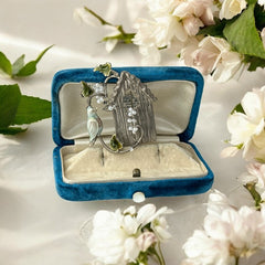 Vintage 1990s silver tone brooch featuring a light blue enamel bird perched on a wire branch with green enamel leaves and white seed bead flowers, set in front of a textured birdhouse; whimsical 3D design in very good vintage condition.