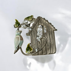 Vintage 1990s silver tone brooch featuring a light blue enamel bird perched on a wire branch with green enamel leaves and white seed bead flowers, set in front of a textured birdhouse; whimsical 3D design in very good vintage condition.
