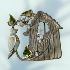 Vintage 1990s silver tone brooch featuring a light blue enamel bird perched on a wire branch with green enamel leaves and white seed bead flowers, set in front of a textured birdhouse; whimsical 3D design in very good vintage condition.
