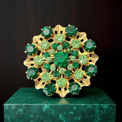 Vintage 1950s gold tone brooch with a ruffled sunburst design, featuring concentric rings of emerald and peridot rhinestones and a large green center stone; bold mid-century costume jewelry with layered sparkle and sculptural detail.