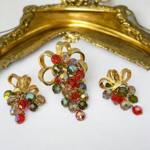 A dazzling vintage 1980s brooch and earrings set featuring faceted AB crystal beads in orange, olivine, and light topaz, forming a grape cluster suspended from a gold tone bow. The aurora borealis finish enhances the sparkle, reminiscent of Juliana-style jewelry. The earrings mirror the brooch’s elegant design, creating a cohesive demi-parure. A timeless statement piece in excellent vintage condition.