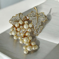 Elegant vintage 1980s brooch featuring a gold-tone grape vine design with round faux pearls forming the grape cluster. The leaves are adorned with sparkling clear pave rhinestones, adding a luxurious touch. Unsigned and in very good vintage condition, this brooch is a timeless statement piece.