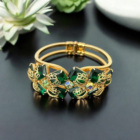 Vintage Juliana clamper bracelet from the 1950s, featuring 10 emerald-shaped rhinestones with filigree leaves, three AB light green rhinestones, and a gold-tone finish. A glamorous statement piece in excellent condition.