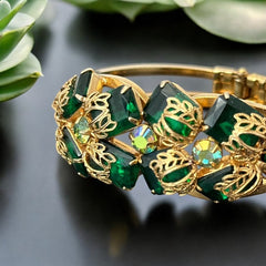 Vintage Juliana clamper bracelet from the 1950s, featuring 10 emerald-shaped rhinestones with filigree leaves, three AB light green rhinestones, and a gold-tone finish. A glamorous statement piece in excellent condition.