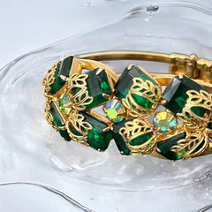 Vintage Juliana clamper bracelet from the 1950s, featuring 10 emerald-shaped rhinestones with filigree leaves, three AB light green rhinestones, and a gold-tone finish. A glamorous statement piece in excellent condition.