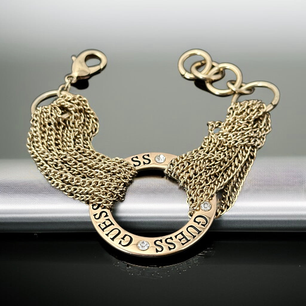 Gold tone Guess bracelet with multi-strand chains and a central ring engraved with ‘GUESS’ and accented by clear rhinestones, shown on neutral backdrop.