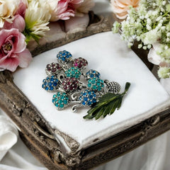 Vintage Hollycraft rhinestone brooch from the 1950s/60s featuring 10 multicolor rhinestone berries in blue, teal green, and dark amethyst hues on silver-tone branches, accented by a green enamel leaf with a small spot of missing enamel at the bottom.