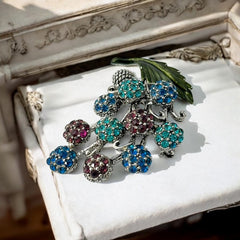 Vintage Hollycraft rhinestone brooch from the 1950s/60s featuring 10 multicolor rhinestone berries in blue, teal green, and dark amethyst hues on silver-tone branches, accented by a green enamel leaf with a small spot of missing enamel at the bottom.