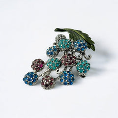 Vintage Hollycraft rhinestone brooch from the 1950s/60s featuring 10 multicolor rhinestone berries in blue, teal green, and dark amethyst hues on silver-tone branches, accented by a green enamel leaf with a small spot of missing enamel at the bottom.
