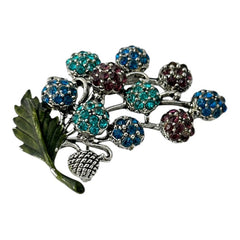 Vintage Hollycraft rhinestone brooch from the 1950s/60s featuring 10 multicolor rhinestone berries in blue, teal green, and dark amethyst hues on silver-tone branches, accented by a green enamel leaf with a small spot of missing enamel at the bottom.