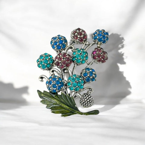 Vintage Hollycraft rhinestone brooch from the 1950s/60s featuring 10 multicolor rhinestone berries in blue, teal green, and dark amethyst hues on silver-tone branches, accented by a green enamel leaf with a small spot of missing enamel at the bottom.