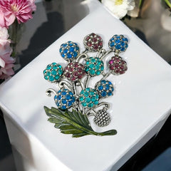 Vintage Hollycraft rhinestone brooch from the 1950s/60s featuring 10 multicolor rhinestone berries in blue, teal green, and dark amethyst hues on silver-tone branches, accented by a green enamel leaf with a small spot of missing enamel at the bottom.