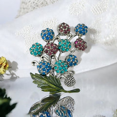 Vintage Hollycraft rhinestone brooch from the 1950s/60s featuring 10 multicolor rhinestone berries in blue, teal green, and dark amethyst hues on silver-tone branches, accented by a green enamel leaf with a small spot of missing enamel at the bottom.