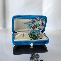 Vintage Hollycraft rhinestone brooch from the 1950s/60s featuring 10 multicolor rhinestone berries in blue, teal green, and dark amethyst hues on silver-tone branches, accented by a green enamel leaf with a small spot of missing enamel at the bottom.