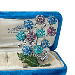Vintage Hollycraft rhinestone brooch from the 1950s/60s featuring 10 multicolor rhinestone berries in blue, teal green, and dark amethyst hues on silver-tone branches, accented by a green enamel leaf with a small spot of missing enamel at the bottom.