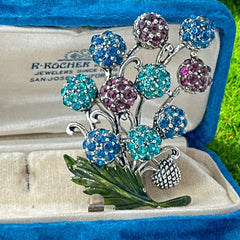 Vintage Hollycraft rhinestone brooch from the 1950s/60s featuring 10 multicolor rhinestone berries in blue, teal green, and dark amethyst hues on silver-tone branches, accented by a green enamel leaf with a small spot of missing enamel at the bottom.