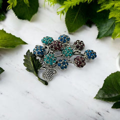 Vintage Hollycraft rhinestone brooch from the 1950s/60s featuring 10 multicolor rhinestone berries in blue, teal green, and dark amethyst hues on silver-tone branches, accented by a green enamel leaf with a small spot of missing enamel at the bottom.