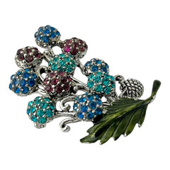 Vintage Hollycraft rhinestone brooch from the 1950s/60s featuring 10 multicolor rhinestone berries in blue, teal green, and dark amethyst hues on silver-tone branches, accented by a green enamel leaf with a small spot of missing enamel at the bottom.