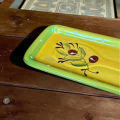 Rectangular ceramic spoon rest with yellow interior and green polka-dot rim, featuring hand-painted olive leaves, red berries, and whimsical fork and spoon motif; measures 11 by 4 inches.
