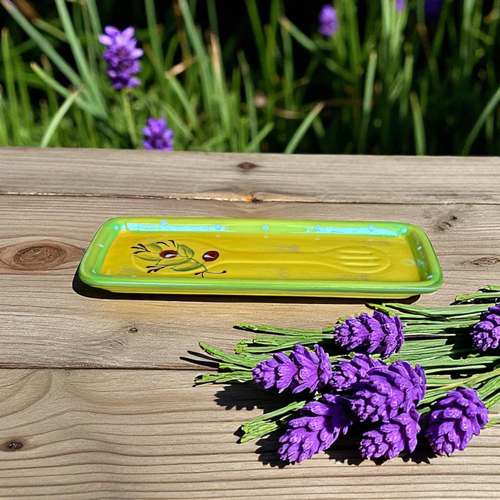 Rectangular ceramic spoon rest with yellow interior and green polka-dot rim, featuring hand-painted olive leaves, red berries, and whimsical fork and spoon motif; measures 11 by 4 inches.