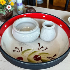Round ceramic serving dish with red and black rim, hand-painted with green olive branches and brown olives on a cream base, accompanied by two small matching vessels nested inside.