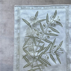 Provence tea towel featuring an olive branch with fruits and flowers at the center, framed by the repeated text 'L'Écomusée de l'Olivier.' Woven in olive green on a natural background with a 100% cotton composition, blending rustic charm and elegance.