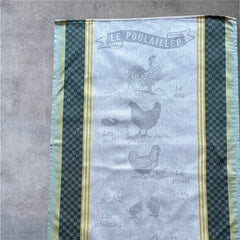 Provence tea towel featuring Le Poulailler design with rooster, hen, eggs, chicks, and chicken motifs. Center in natural and light gray tones, bordered with yellow stripes, dark green checkered pattern, and lighter green edges. Made of 100% cotton, ideal for French farmyard decor.
