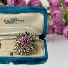 Vintage sunburst brooch with pink and clear rhinestones set in gold tone metal, featuring pointed petal-like rays and a central cluster of pink stones, photographed front and back to highlight its 1980s design and secure clasp.