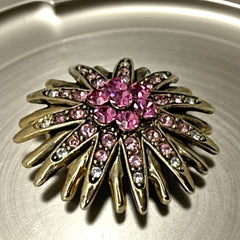 Vintage sunburst brooch with pink and clear rhinestones set in gold tone metal, featuring pointed petal-like rays and a central cluster of pink stones, photographed front and back to highlight its 1980s design and secure clasp.