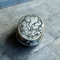 A vintage Tibetan 'silver' and turquoise bead from the 1990s, featuring a turquoise-green oval cabochon with natural matrix, set in a handcrafted bezel. The back of the bead displays intricate repousse work with swirling cloud and flower designs, iconic in Tibetan jewelry.