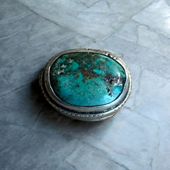 A vintage Tibetan 'silver' and turquoise bead from the 1990s, featuring a turquoise-green oval cabochon with natural matrix, set in a handcrafted bezel. The back of the bead displays intricate repousse work with swirling cloud and flower designs, iconic in Tibetan jewelry.