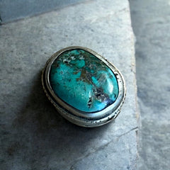 A vintage Tibetan 'silver' and turquoise bead from the 1990s, featuring a turquoise-green oval cabochon with natural matrix, set in a handcrafted bezel. The back of the bead displays intricate repousse work with swirling cloud and flower designs, iconic in Tibetan jewelry.