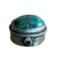 A vintage Tibetan 'silver' and turquoise bead from the 1990s, featuring a turquoise-green oval cabochon with natural matrix, set in a handcrafted bezel. The back of the bead displays intricate repousse work with swirling cloud and flower designs, iconic in Tibetan jewelry.
