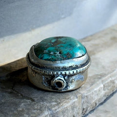 A vintage Tibetan 'silver' and turquoise bead from the 1990s, featuring a turquoise-green oval cabochon with natural matrix, set in a handcrafted bezel. The back of the bead displays intricate repousse work with swirling cloud and flower designs, iconic in Tibetan jewelry.