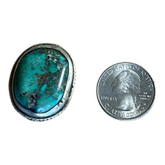 A vintage Tibetan 'silver' and turquoise bead from the 1990s, featuring a turquoise-green oval cabochon with natural matrix, set in a handcrafted bezel. The back of the bead displays intricate repousse work with swirling cloud and flower designs, iconic in Tibetan jewelry.