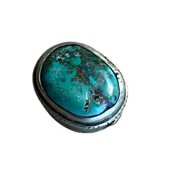 A vintage Tibetan 'silver' and turquoise bead from the 1990s, featuring a turquoise-green oval cabochon with natural matrix, set in a handcrafted bezel. The back of the bead displays intricate repousse work with swirling cloud and flower designs, iconic in Tibetan jewelry.