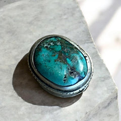 A vintage Tibetan 'silver' and turquoise bead from the 1990s, featuring a turquoise-green oval cabochon with natural matrix, set in a handcrafted bezel. The back of the bead displays intricate repousse work with swirling cloud and flower designs, iconic in Tibetan jewelry.