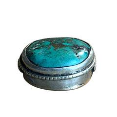 A vintage Tibetan 'silver' and turquoise bead from the 1990s, featuring a turquoise-green oval cabochon with natural matrix, set in a handcrafted bezel. The back of the bead displays intricate repousse work with swirling cloud and flower designs, iconic in Tibetan jewelry.