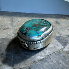 A vintage Tibetan 'silver' and turquoise bead from the 1990s, featuring a turquoise-green oval cabochon with natural matrix, set in a handcrafted bezel. The back of the bead displays intricate repousse work with swirling cloud and flower designs, iconic in Tibetan jewelry.