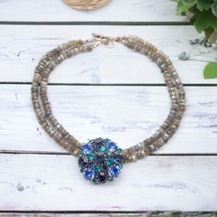 A stunning vintage Weiss crystal dome brooch in shades of blue and teal, paired with a double strand of shiny blue and gray faceted labradorite rondelles. The brooch can be worn centered, off-center, or separately, offering versatile styling. Finished with sterling silver spacers and a toggle clasp, this unique piece blends vintage charm with modern craftsmanship, signed Weiss on the back and featuring the EM tag in Etruscan alphabet for Elisabeth Michel-Meyrueix.